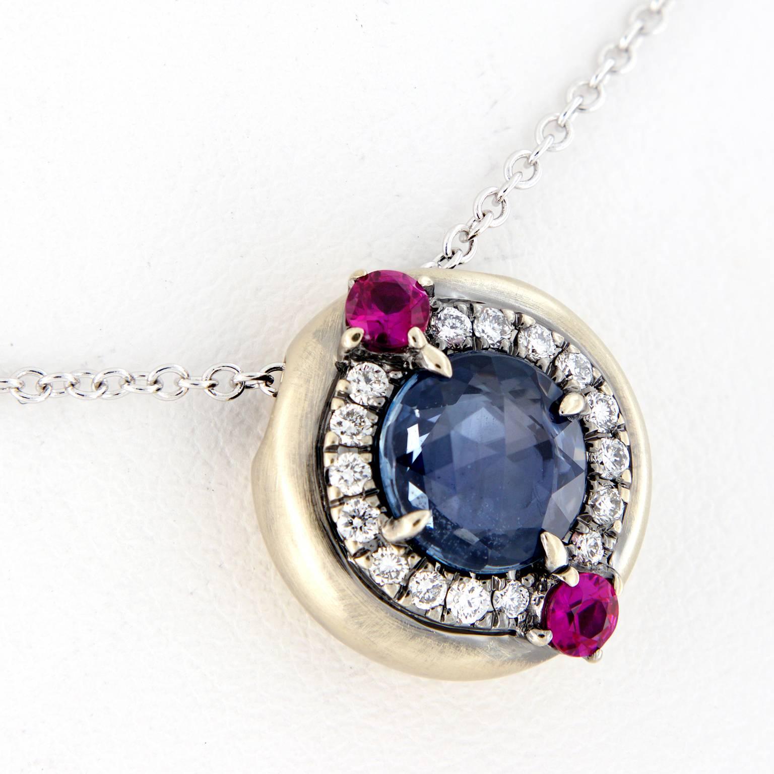 Danhier’s Galaxy pendant necklace from the Evolution Collection centers around a round rose-cut blue sapphire, enhanced with a halo of white diamonds and two rubies. 18 inch chain is 18k white gold and pendant features a hand-rubbed patina. Pendant
