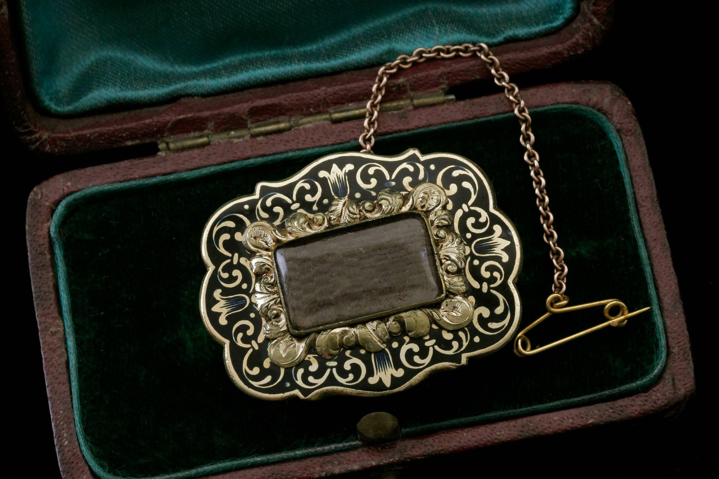 C.1820-1830.  A late Georgian or early Victorian mourning brooch. The 15k gold cased mount is beautifully adorned with black enamel. The center locket, containing woven hair, is decorated with highly raised, gold leafy scrolls around its frame. It