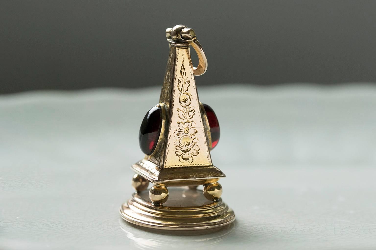 C.1870. An unusual 18k gold obelisk seal fob. The fob is in the shape of obelisk, and two garnet cabochons adorn two sides of it. The seal is made of chalcedony, and the carved motto says in French 'Je Ne Change Qu'en Mourant'; 'I change only in