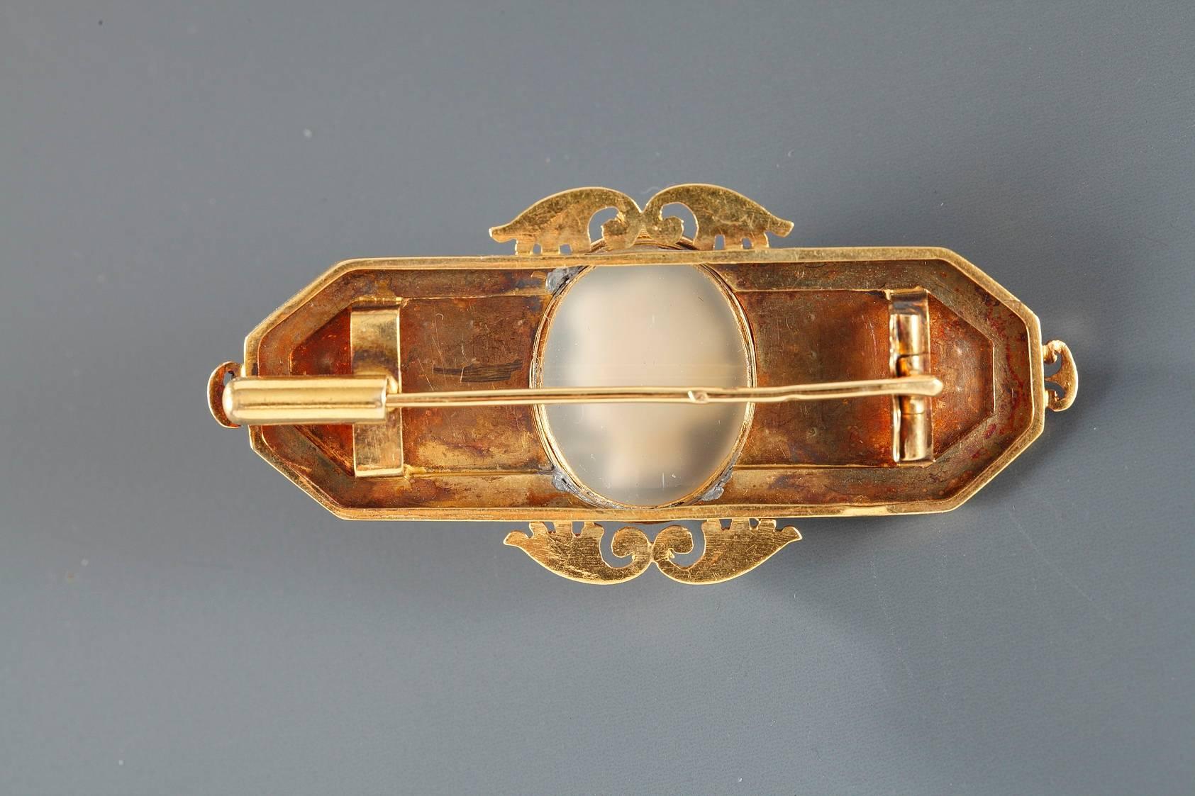 Gold brooch featuring a double-layered agate cameo of a woman. Openwork palmettes set off the top and the bottom of the cameo, which is set on a gold, rectangular background. The background is delicately sculpted with flowers separated by rounded