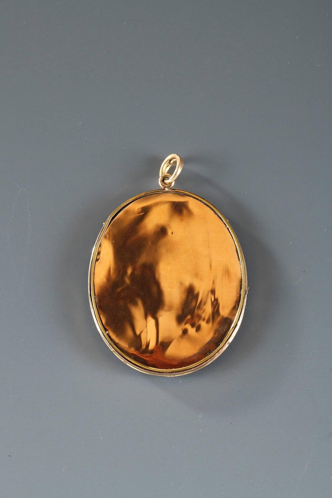 Flat pendant in the 18th century pastoral style, showing a young woman carrying roses in her gathered skirt and walking toward her suitor, who is sitting on the grass. A castle is visible in the background. It is mounted in gold with a black enamel