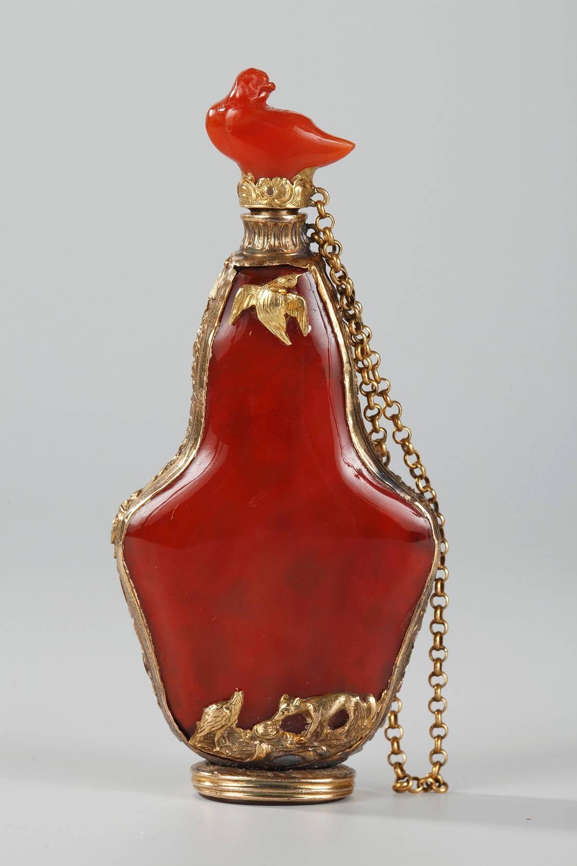 Pear-shaped flask in orange-red carnelian gemstone, set in a gold mounting that is intricately embellished with stylized flowers. Each side of the mounting’s base features two animals: on one side, a fox is watching a stork with its beak in a vase,
