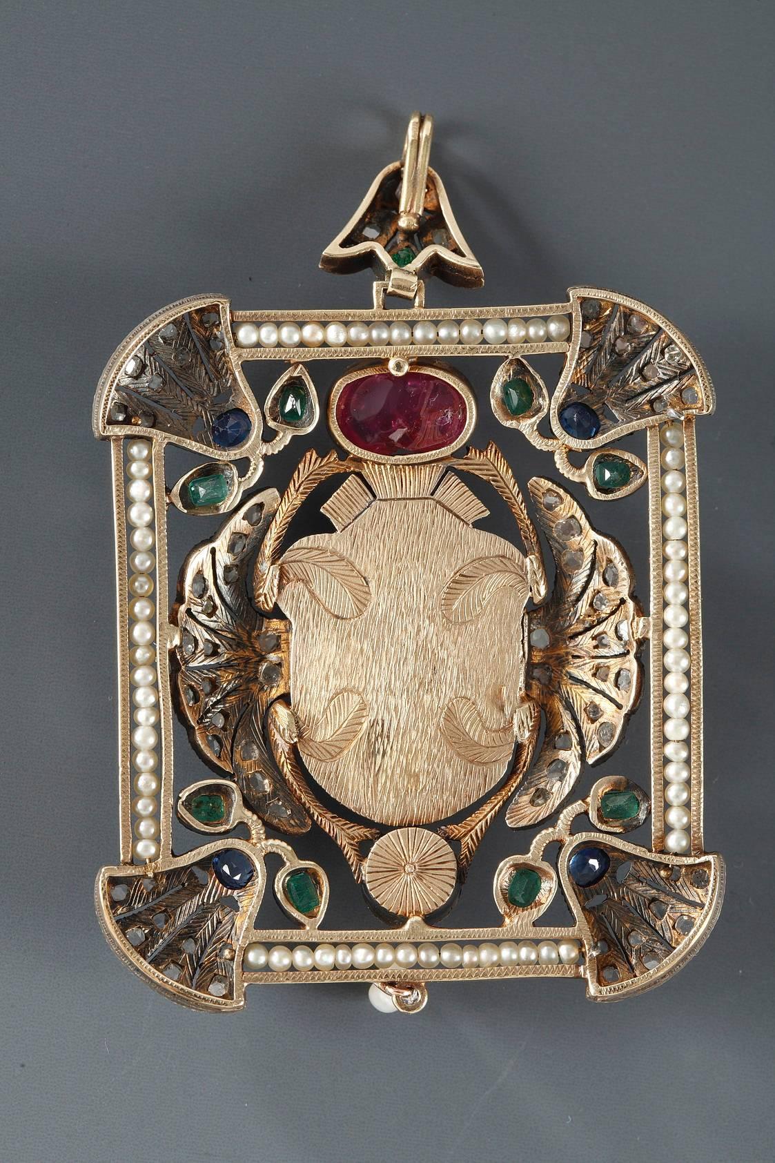 Silver and 14k gold-mounted pendant featuring a winged scarab beetle. The body of the beetle is decorated with square-cut emeralds, and a large, ruby cabochon is set above its head. It is surrounded with a floral decoration that is highlighted with