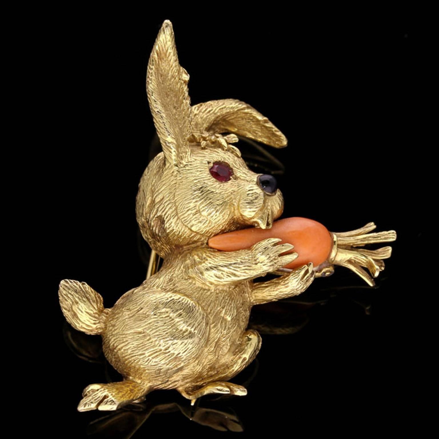 The delightful little rabbit finely crafted from textured 18ct yellow gold, one ear pointing straight up, the other flopping down whilst he busily munches on a carrot carved from coral held in his front paws, his eyes are round brilliant rubies and