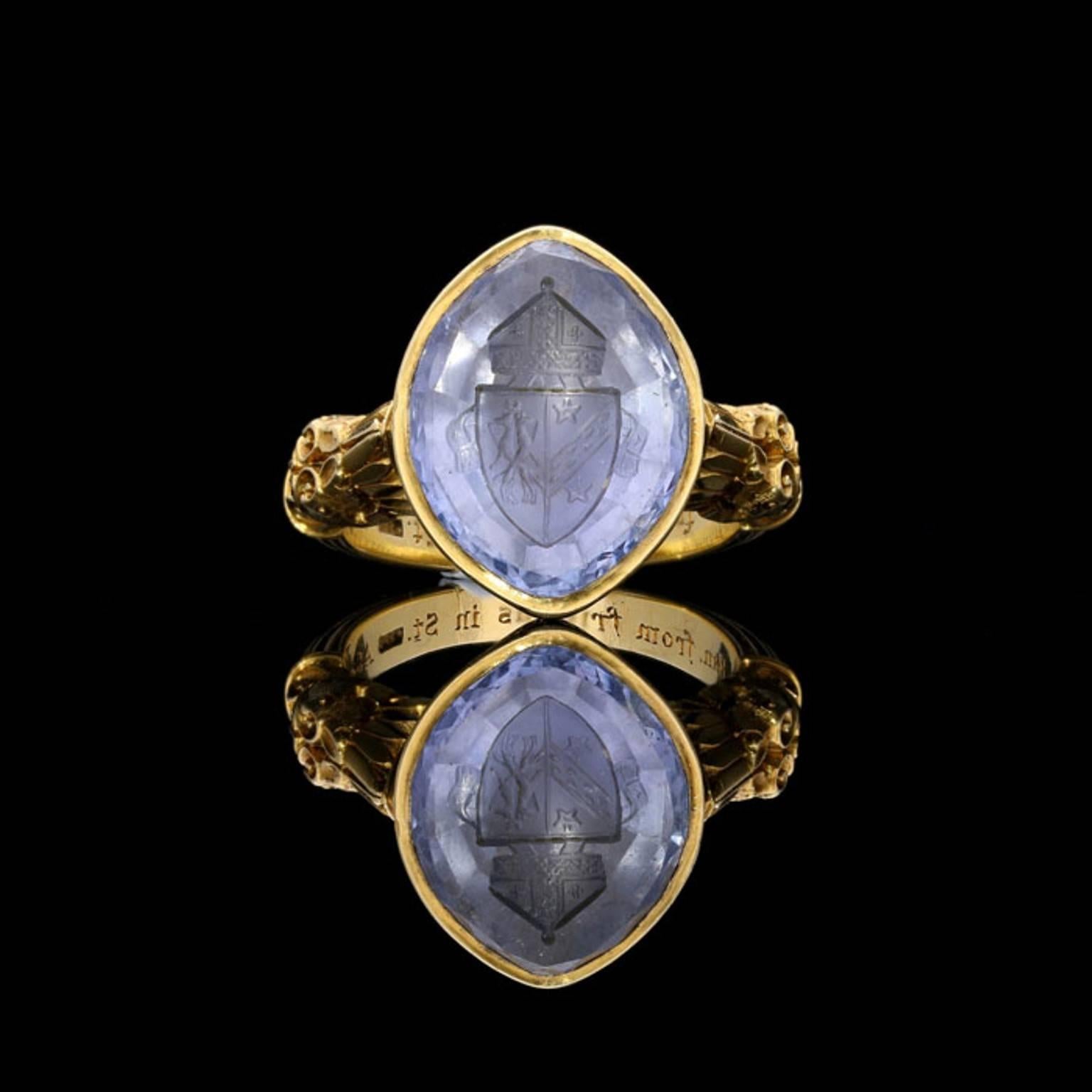 A highly decorative and ornate Bishop's ring carat 1899, finely crafted in 18 carat yellow gold with a large marquise-shaped pale blue sapphire intaglio carved with a mitre atop a shield depicting the coat of arms for Bangor coupled with that of