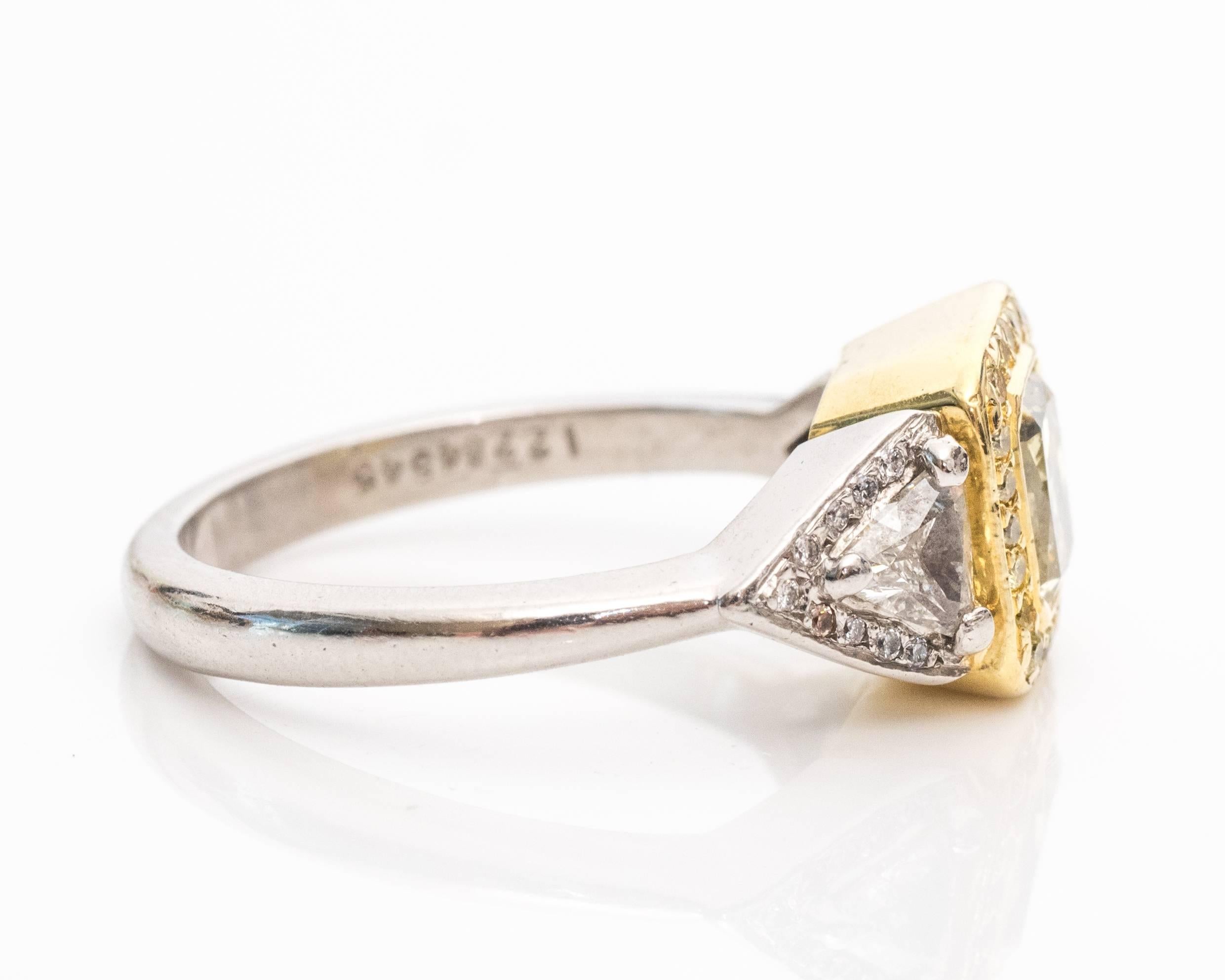 The radiant cut center diamond has official certification from 2005. 
The color is a distinct fancy yellow hue with a halo of small fancy yellow diamonds surrounding the center stone. This area of the stone is mounted in 18 karat yellow gold to