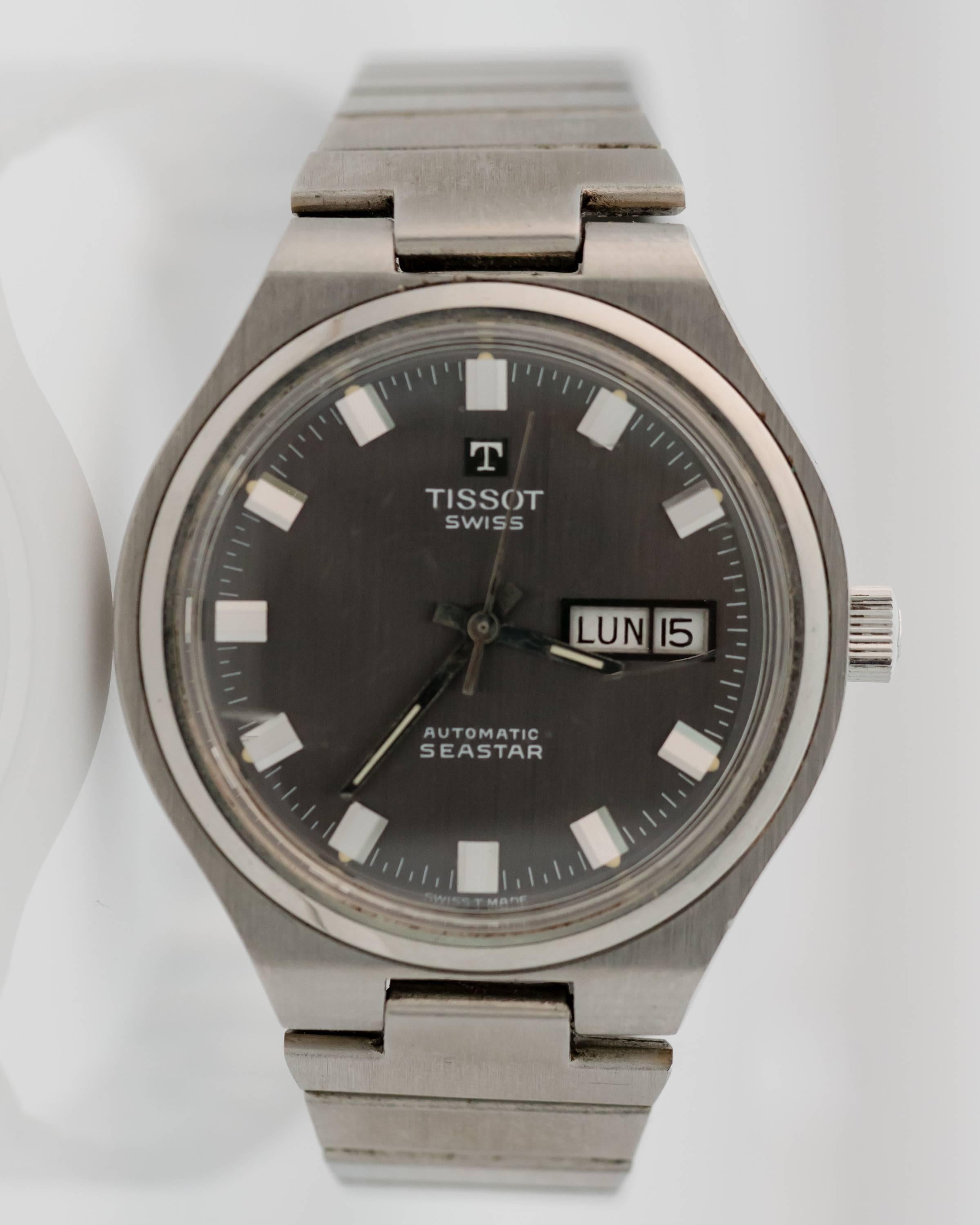 This 1955 Retro Tissot Automatic Stainless Steel Seastar watch features a Spanish language day and date window, slate grey stick dial, acrylic crystal, lightly brushed stainless steel case and stainless steel bracelet with deployment clasp. The