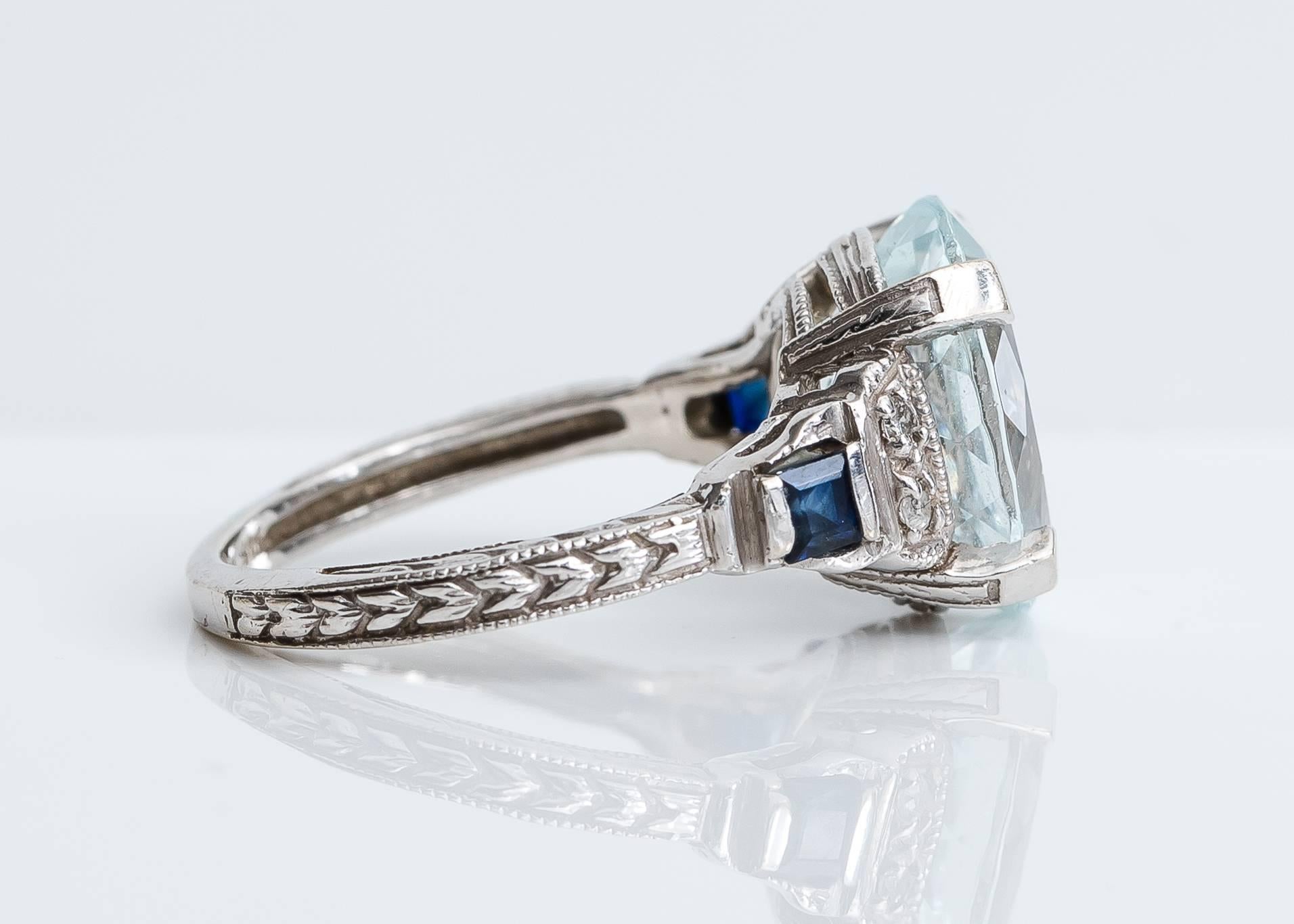 This stunning 1950s Art Nouveau-inspired 3 Carat Aquamarine Ring features a center stone flanked by round brilliant diamonds and square French cut sapphires prong set in 14K White Gold. Delicate patterns adorn the top, bottom and sides of the ring