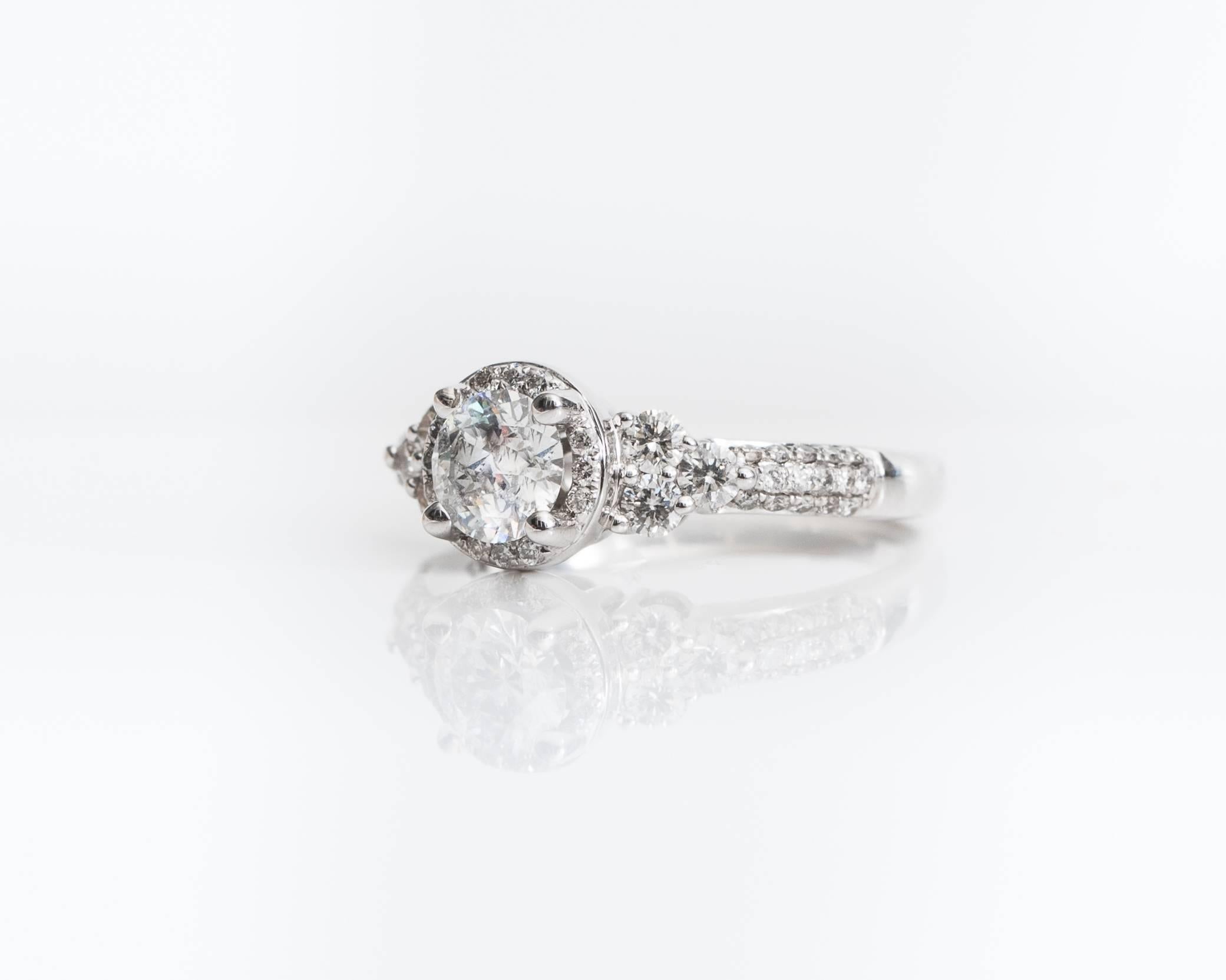 This gorgeous Diamond Engagement Ring features a .75 Carat Round Brilliant Diamond prong set in a 14 Karat White Gold Cathedral Mounting. The center stone is surrounded by a Halo of round brilliant diamonds. The front half of the ring shank is set