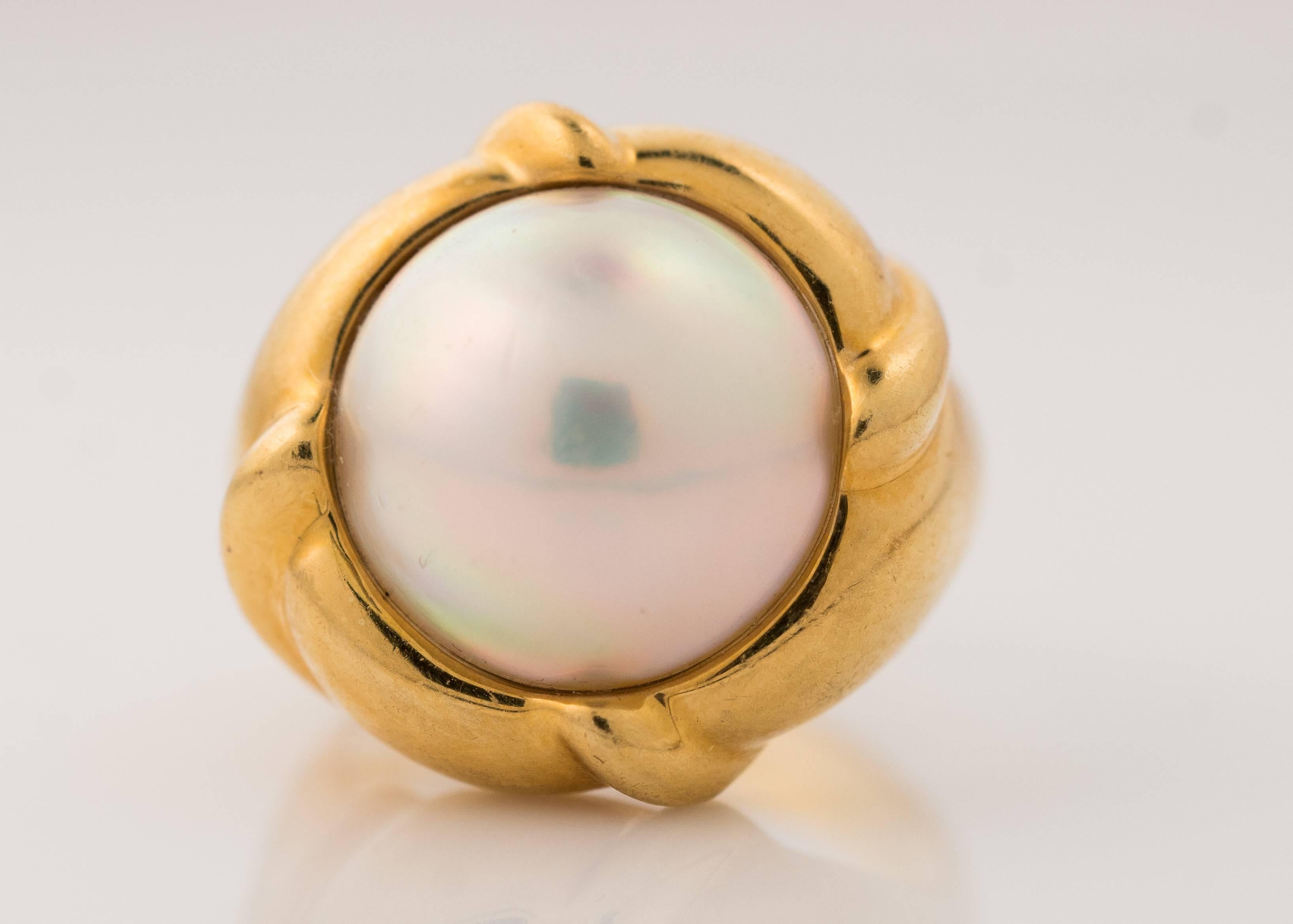 1980s Tiffany and Co. Pearl Cocktail Ring. 
Features a 13.5 millimeter Mabe Pearl in 18 Karat Yellow Gold. This gorgeous, opalescent Pearl is rich with pink, purple and blue hues. Four smooth ribbons of gold wrap from the inner edge of the band to