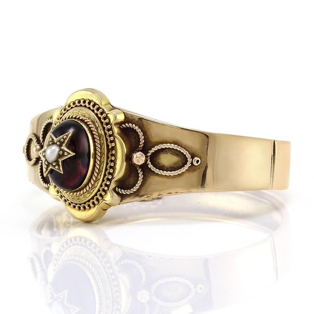 Antique Victorian garnet and pearl hinged bangle bracelet in 15K yellow gold. There are one oval garnet cabochon (9.50ct) and one off-round pearl (4.0mm x 3.0mm). The garnet is bezel set, and the pearl is bead set on a satellite of gold affixed to