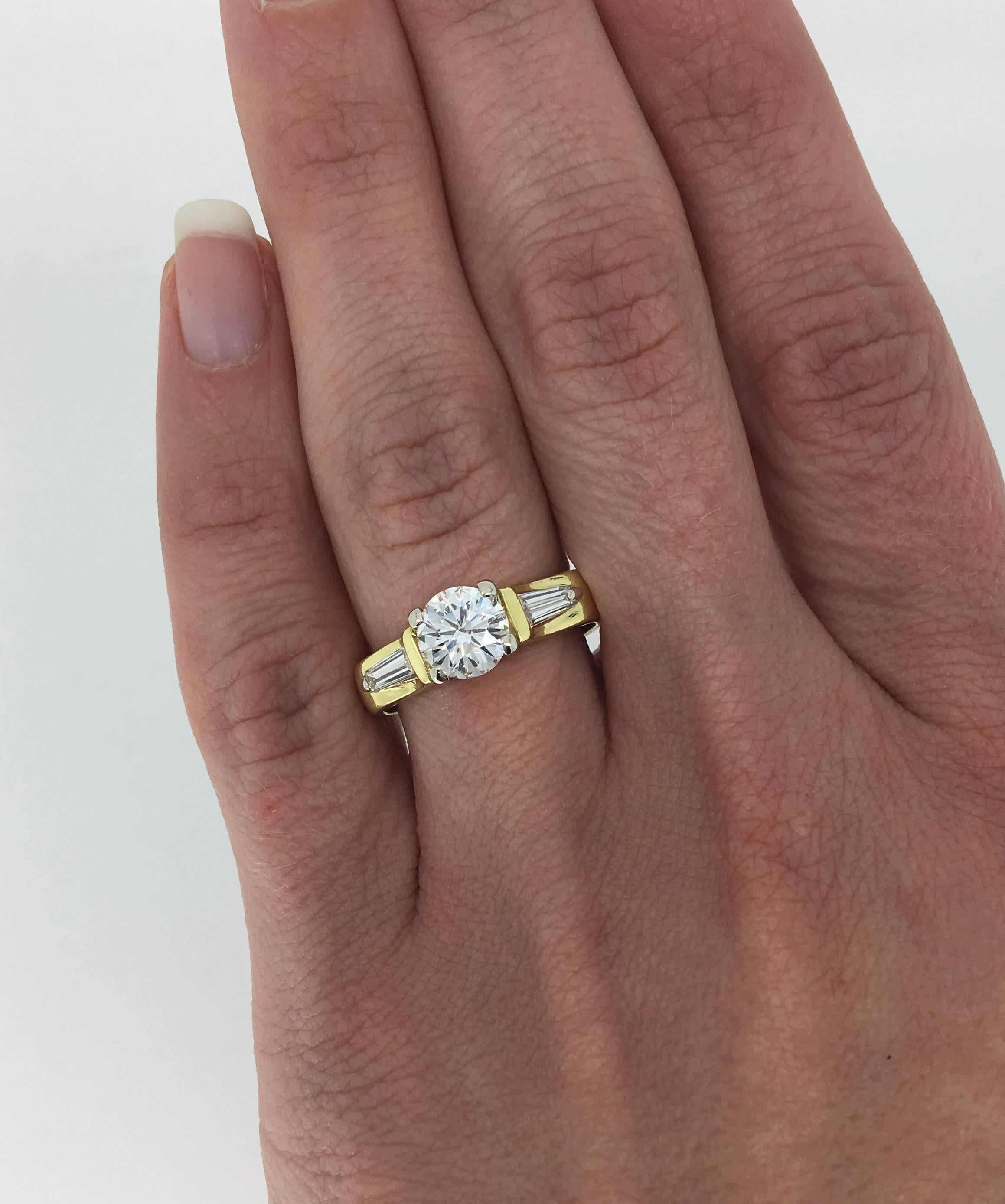 Custom 18K yellow gold engagement ring features a 1.05CT Round Brilliant Cut Diamond that displays G color and SI2 clarity. There are two Tapered Baguette Cut Diamonds accenting the featured Diamond. This ring houses approximately 1.29CTW of