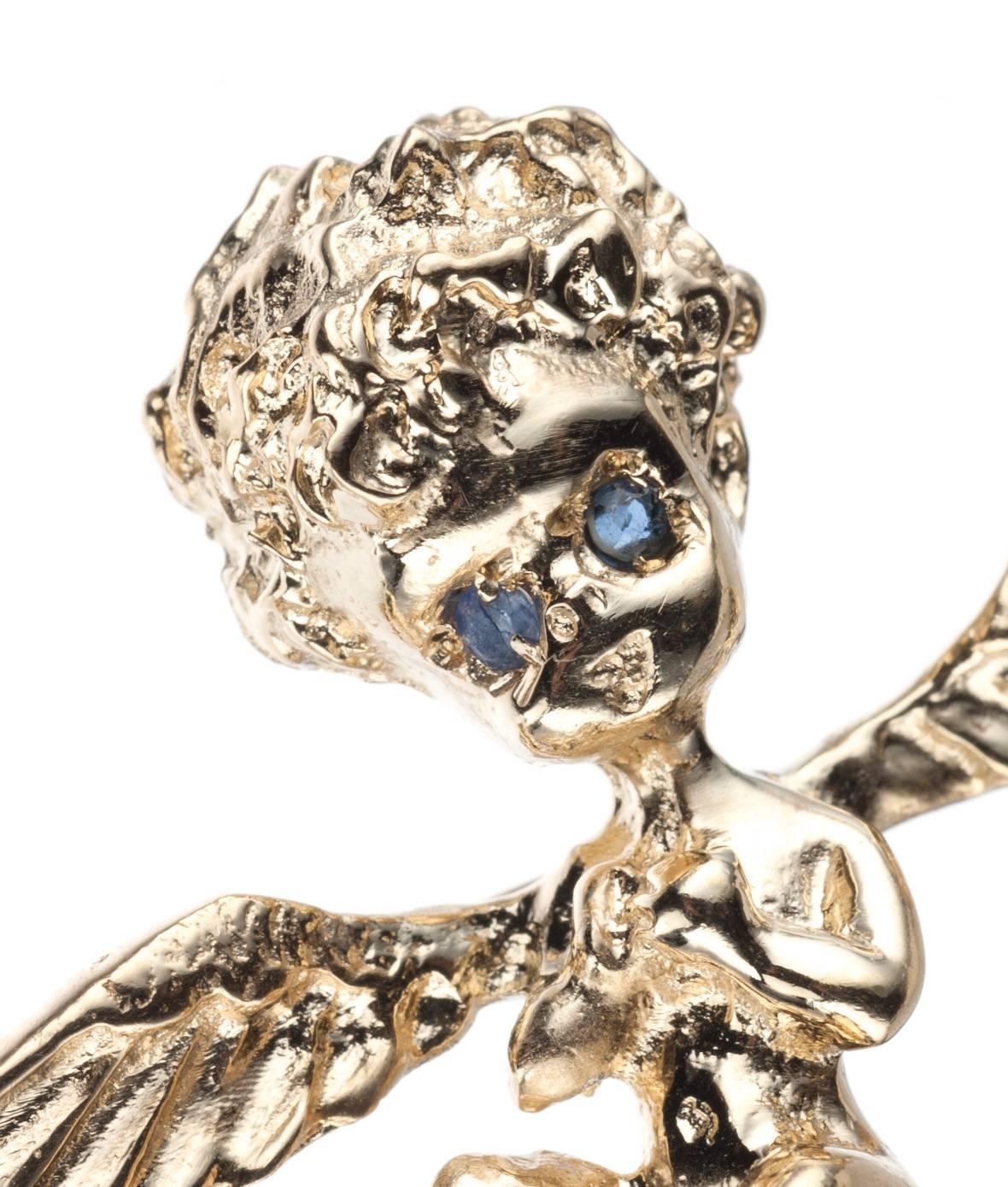 Made in the style of Ruser, a 14-karat yellow gold cherub brooch. The angel rest atop a cloud-shaped baroque cultured pearl, approx. 0.33” wide, and has two round blue sapphires for eyes. Fastens with pin stem and catch. Measures approx. 1.5” long