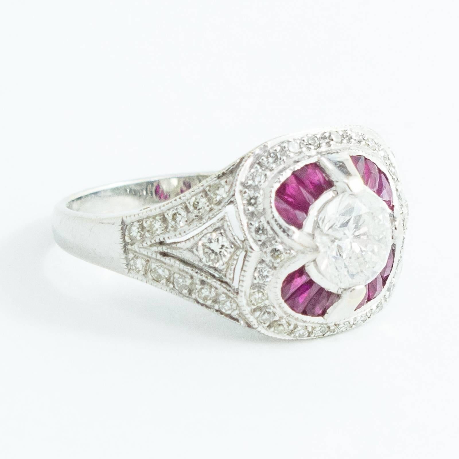A stunning ring is center set with a .72 carat round, brilliant cut Diamond surrounded by 12 calibre cut tapered baguette natural Red Rubies (Corundums) totaling .55 carat and further embellished with 48 round, brilliant cut Diamonds. This 18K white