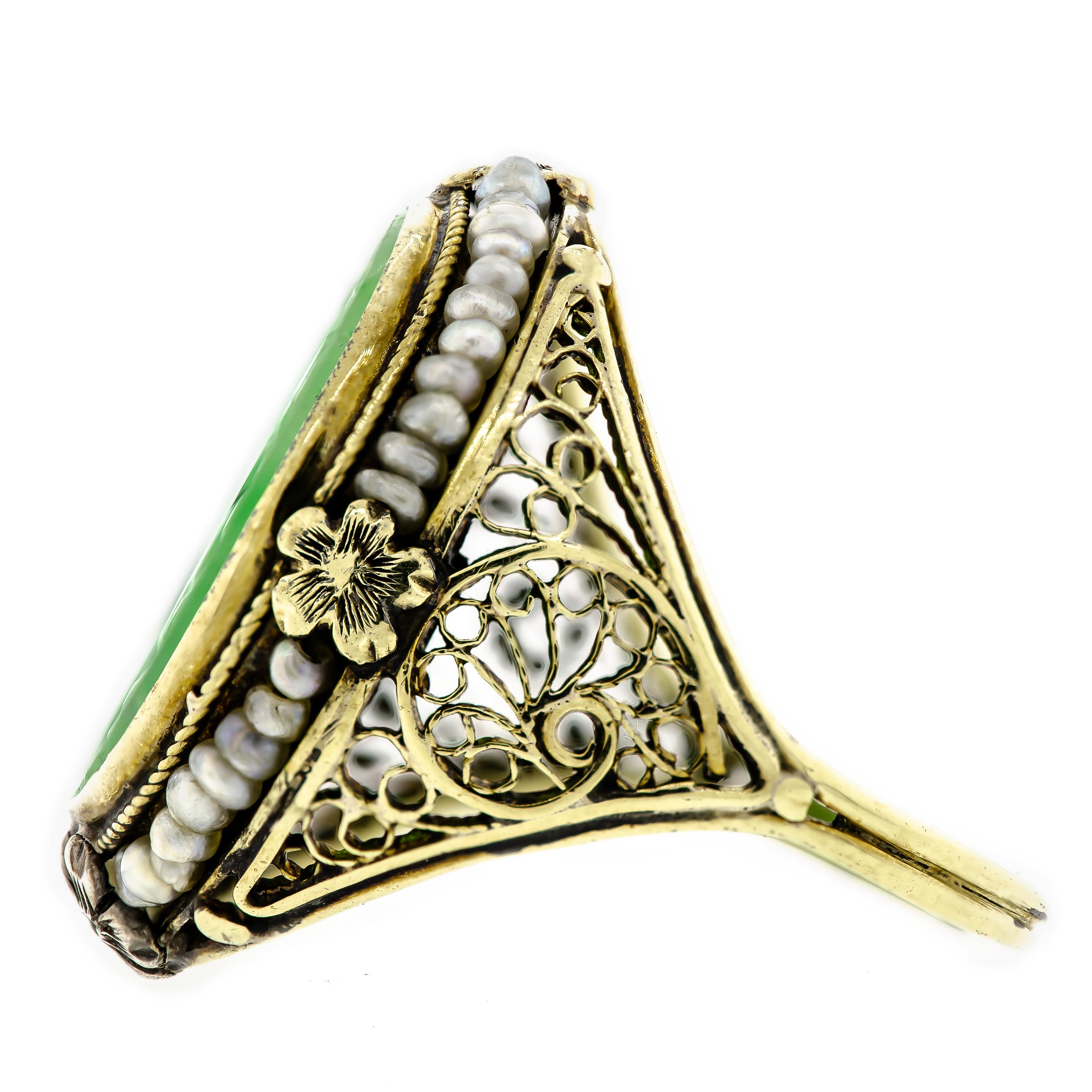 Lovely jadeite, and seed pearl Art Deco ring handcrafted in 14kt yellow gold depicting an elegant foliate design on the mount with beautiful filigree wirework. The center stone is engraved jade featuring a soft foliate design on the jadeite tablet