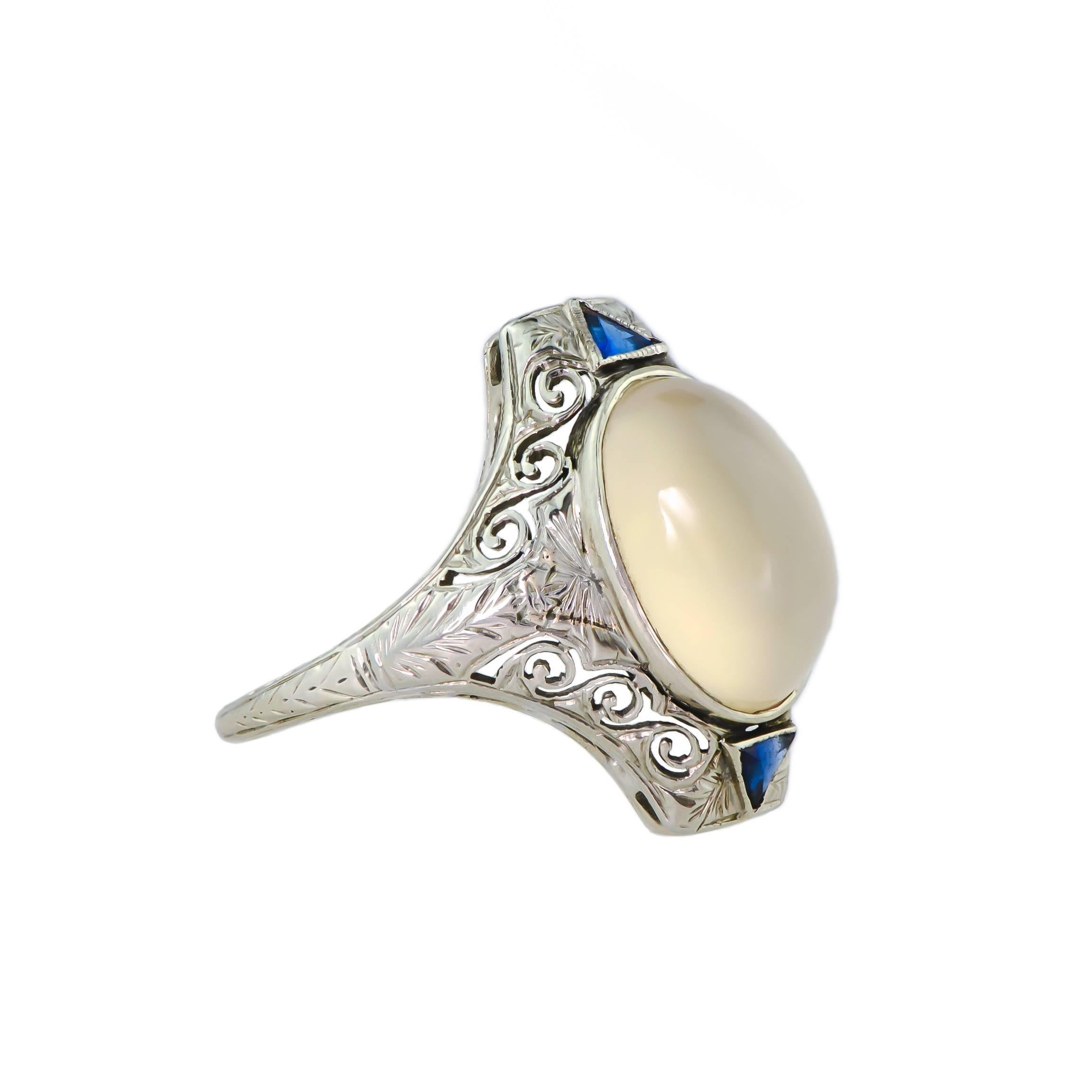 Art Deco circa 1920 moonstone and 14kt white gold filigree ring containing one oval moonstone measuring approximately 13.5mm by 9.5mm bezel set into a pierced and filigree 14kt white gold ring mount further accented with two triangular cut synthetic