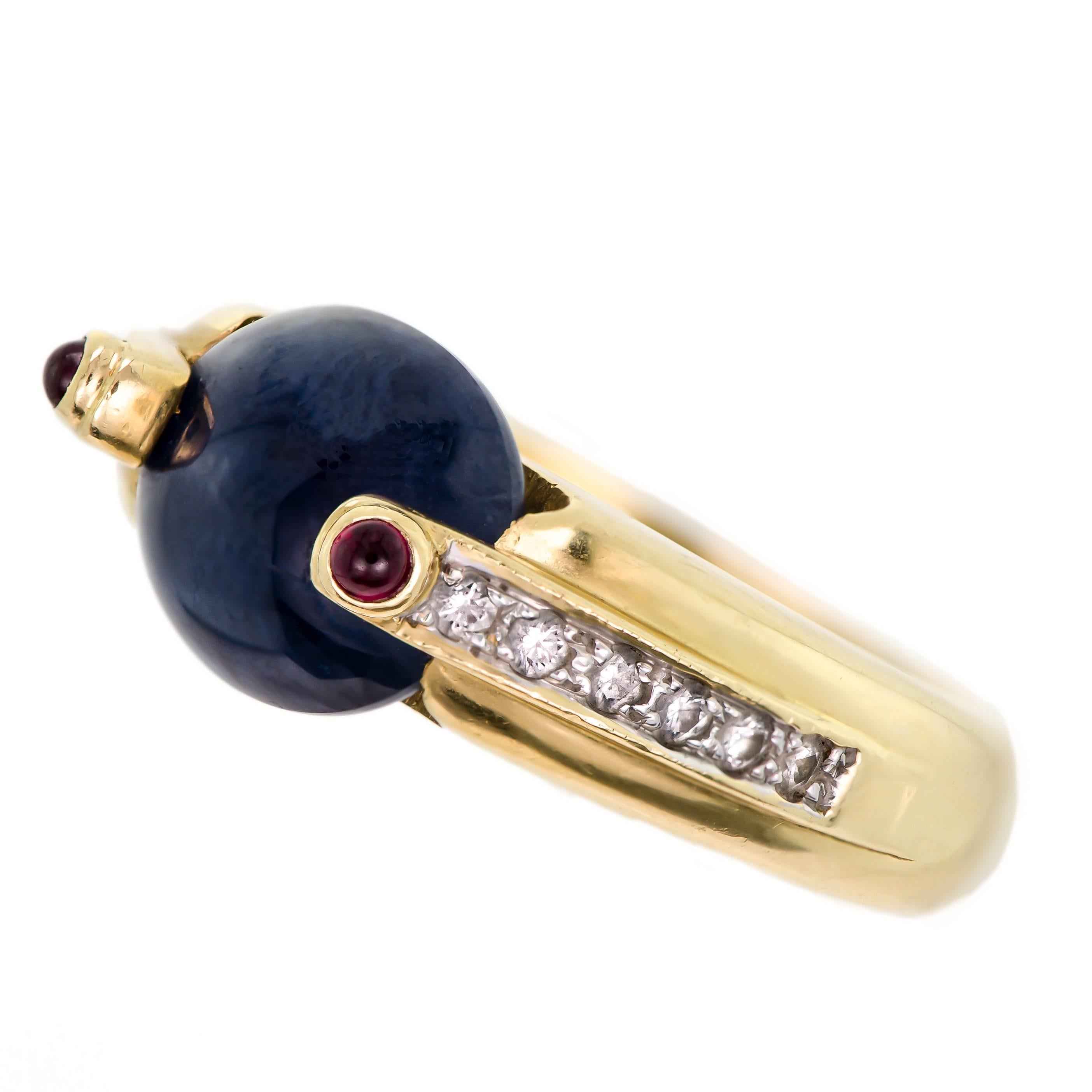 Contemporary 18kt (750 stamp) yellow gold, European sapphire, diamond, and ruby ring containing one (1) oval sapphire cabochon set lengthwise measuring approximately 11mm by 9.5mm accented by twelve (12) (6 each side) small brilliant cut diamond and