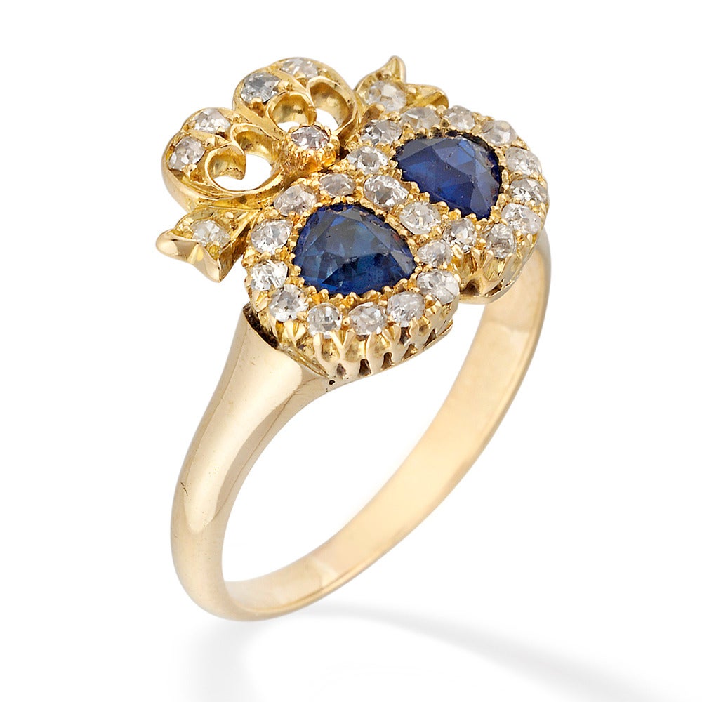 A Victorian sapphire and diamond twin heart ring, each heart with a pear shaped faceted sapphire, weighing approximately 0.40 carats, surrounded by an old brilliant-cut diamond cluster, surmounted by diamond-set ribbon bow, all claw set to gold