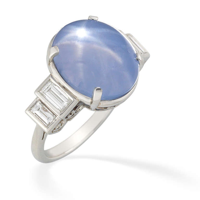 A star sapphire and diamond ring, the pale blue star sapphire weighing 17.70 carats four claw set between two stepped baguette-cut diamonds to each shoulder, the diamonds weighing a total of 0.77 carats, all set in platinum to an open scroll gallery