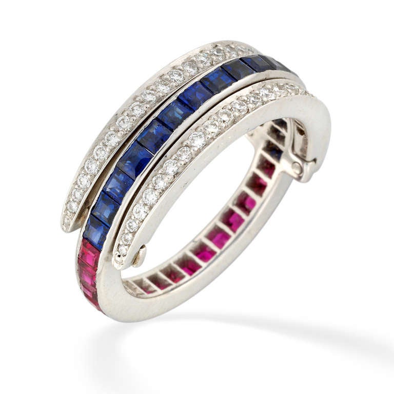 A sapphire, ruby and diamond swivel ring, with a row of french-cut channel-set sapphires, framed by two bands of graduating round brilliant-cut diamonds that pivot to frame an alternate row of french-cut channel-set rubies, all set in millegrain