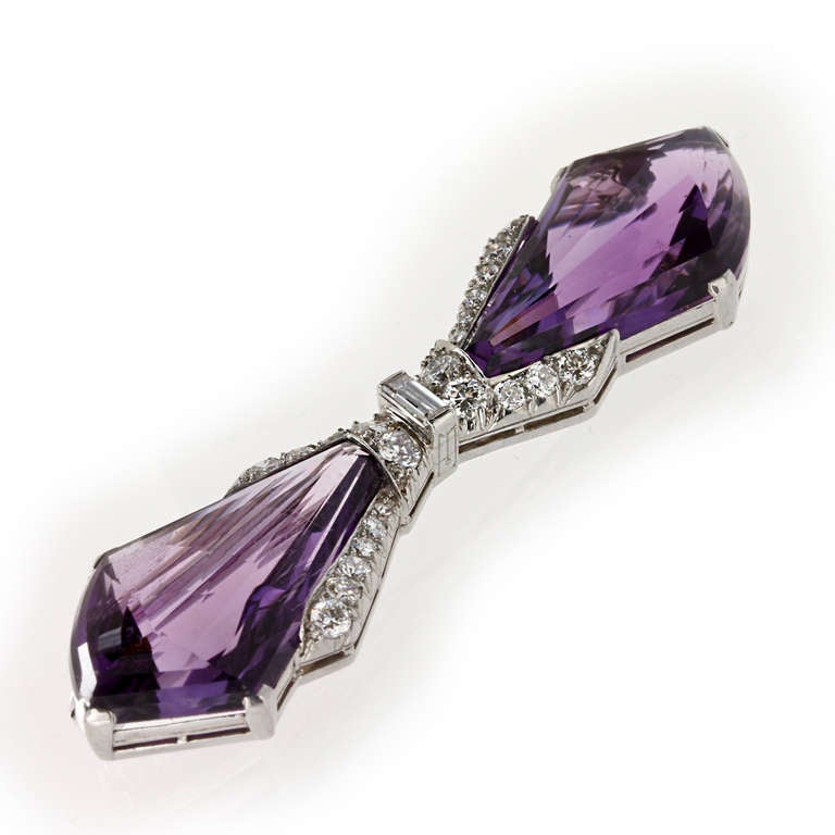 An Art Deco amethyst and diamond bow brooch, the modified kite shaped faceted amethyst bows, each estimated to weigh 7 carats each, claw set to a platinum mount, to the centre a rubover set baguette-cut diamond estimated to weigh 0.07 carats, to