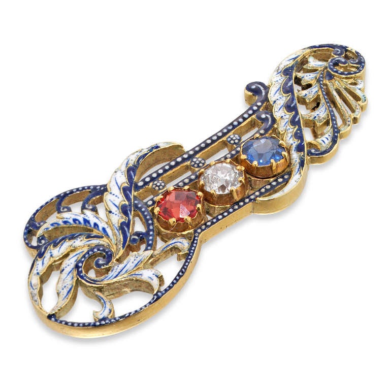 A sapphire, ruby, diamond and enamel brooch by Giuliano, with a central round brilliant-cut diamond, flanked by a round faceted ruby to one side  and a round faceted sapphire to the other, all gems set within a decorative scrolled openwork blue and