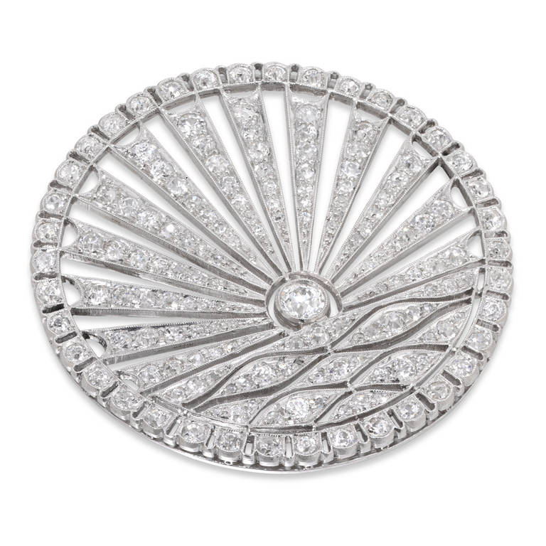 An Art Deco diamond brooch, the circular plaque depicting a stylised rising sun over water, the lower section of undulating form with an old brilliant-cut diamond 'sun' above, within an openwork radiating frame, to an outer scalloped border, white