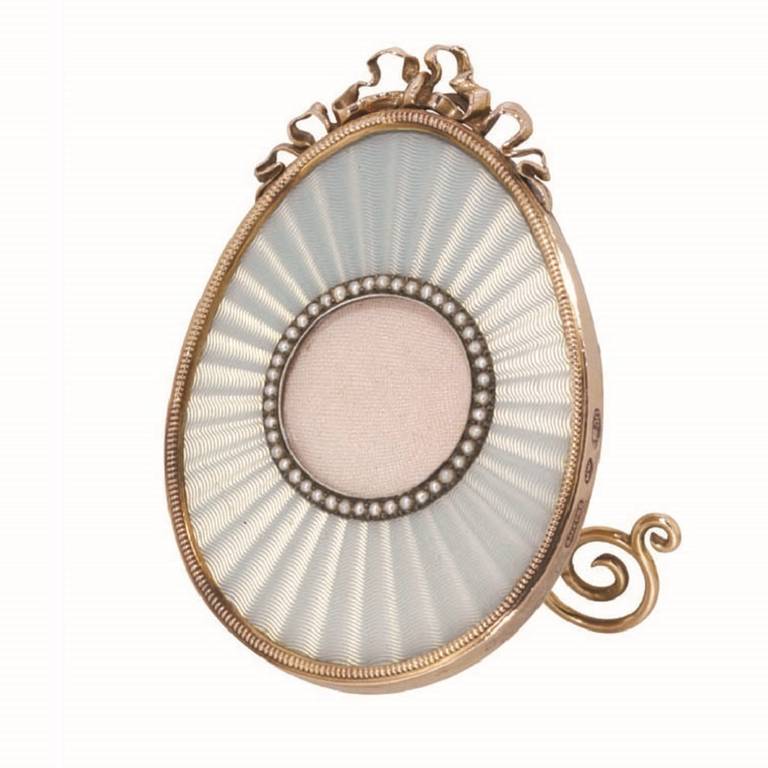 A Fabergé white enamel egg shaped miniature frame, the translucent enamel on an engine turned radiating guilloché ground, with a picture aperture surrounded by 40 seed pearls in a silver setting, the yellow gold outer edge with a ribbon and bow