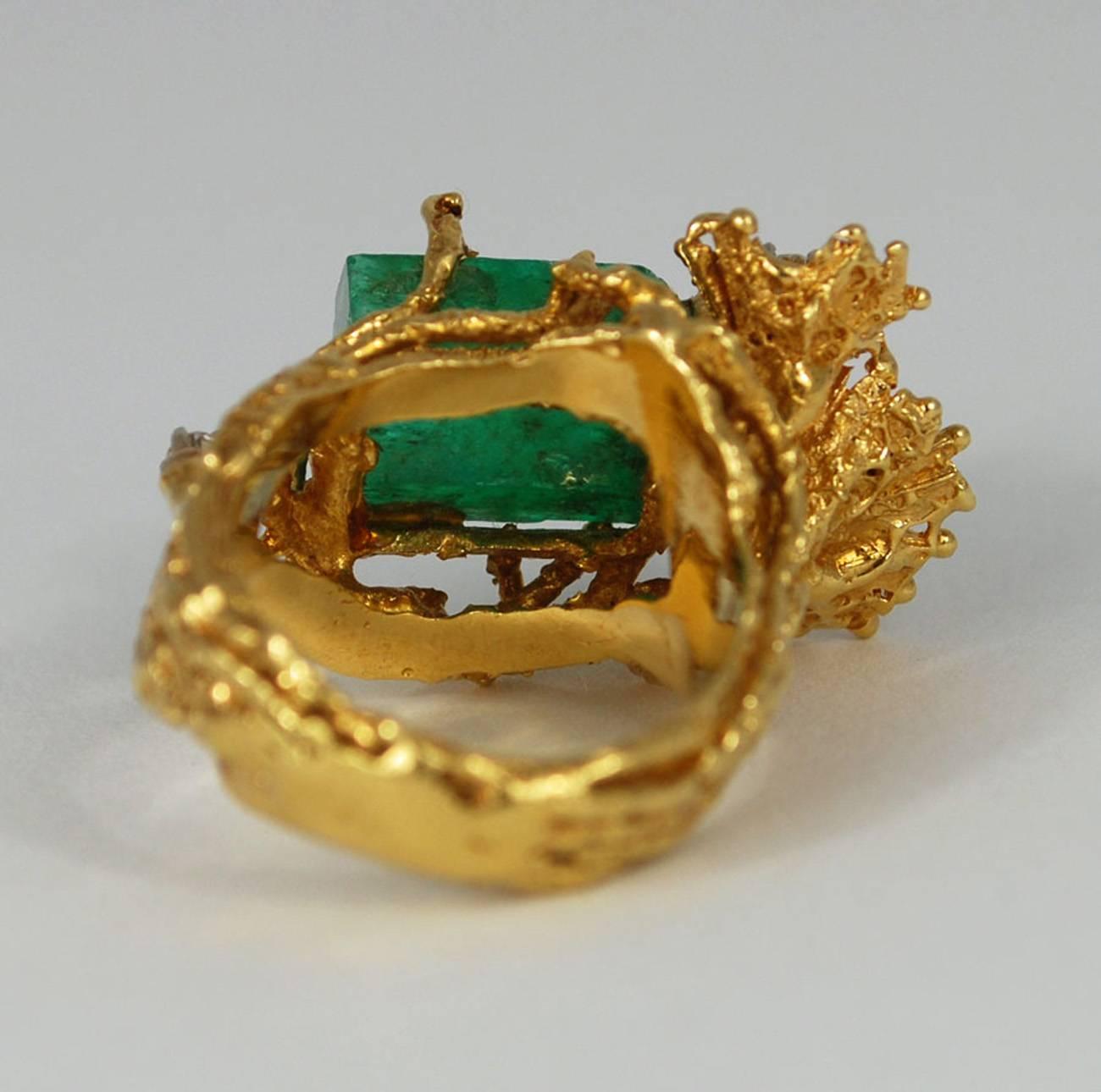 A thick, naturalistic 18K gold band splits into branches wrap around to hold in a large raw emerald stone. At its side, two round cut diamonds are set into gold abstract flowers. A third diamond adds sparkle to the other side of the emerald. Marked