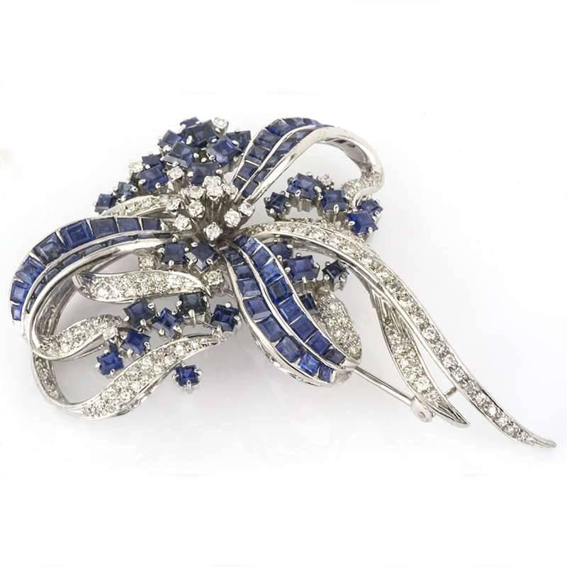 An elegant 14k white gold diamond and sapphire brooch. The central cluster of round cut diamonds are surrounded by a triple sapphire bow and diamond set ribbons. The brooch is made up of square cut sapphires and old cut diamonds, diamonds totalling