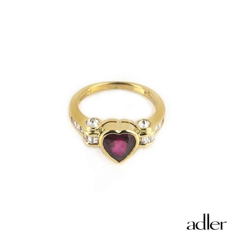 An 18k yellow gold ruby and diamond ring by Adler. The ring is set to the centre with a heart shape faceted ruby weighing approximately 1.45ct displaying a beautiful rich even tone. The heart is set within an elaborate 18k yellow gold graduated