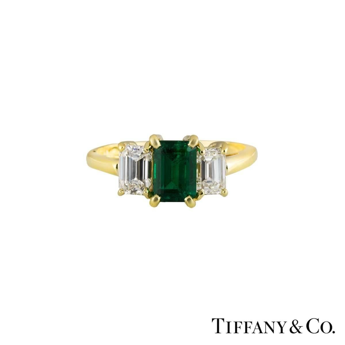 A luxurious 18k Tiffany & Co. diamond and emerald trilogy ring. The ring comprises of an emerald cut emerald set to the centre in a 4 claw setting, with a total approximate weight of 0.75ct and a deep green hue throughout. There are 2 emerald cut