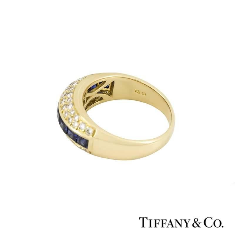 A gorgeous 18k yellow gold dress ring by Tiffany & Co. The ring is horizontally set through the centre with princess cut sapphires, each displaying a deep blue hue, totalling approximately 0.80ct. Accentuating the sapphires are pave set round