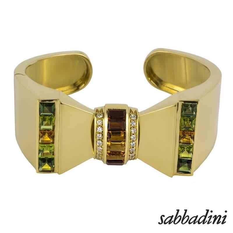 A contemporary 18k yellow gold gem set bangle by Sabbadini. The bangle is composed of a bow motif, set to the centre with rectangular cut citrines, ranging in a hue of deep to light oranges, complemented by a round brilliant cut diamond boarder. The