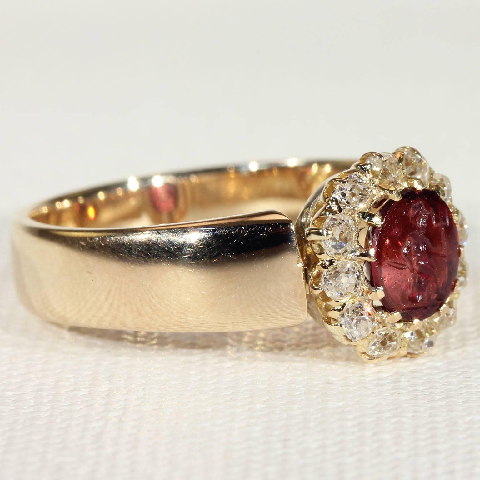 A cluster of Old European cut diamonds surround a carved intaglio garnet on this Victorian Era ring. Handcrafted and marked for the year 1900, this piece features a motif of a man with his pitchfork, which has been carved into the red garnet kissed