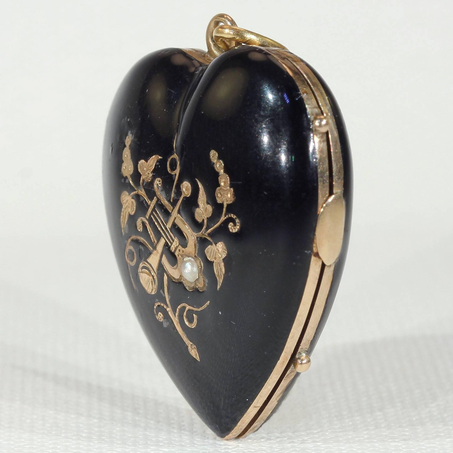 This antique Victorian black enameled and gold heart mourning locket was handcrafted around 1870 in England. The front features a golden trumpet and lyre, surrounded by an array of golden flowers. A 2 mm diameter pearl decorates the stringed