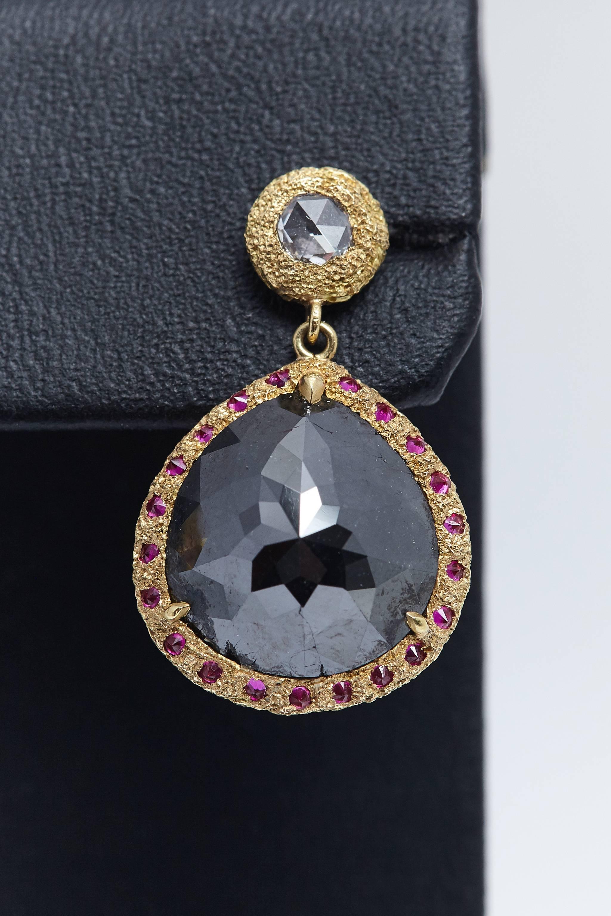 Pair of Treated Black Rose Cut Diamonds with a total weight of approximately 10.30 carats. The diamonds are encircled by rubies which are mounted in eighteen karat yellow gold earrings. The larger diamonds are dangling from a pair of white rose cut