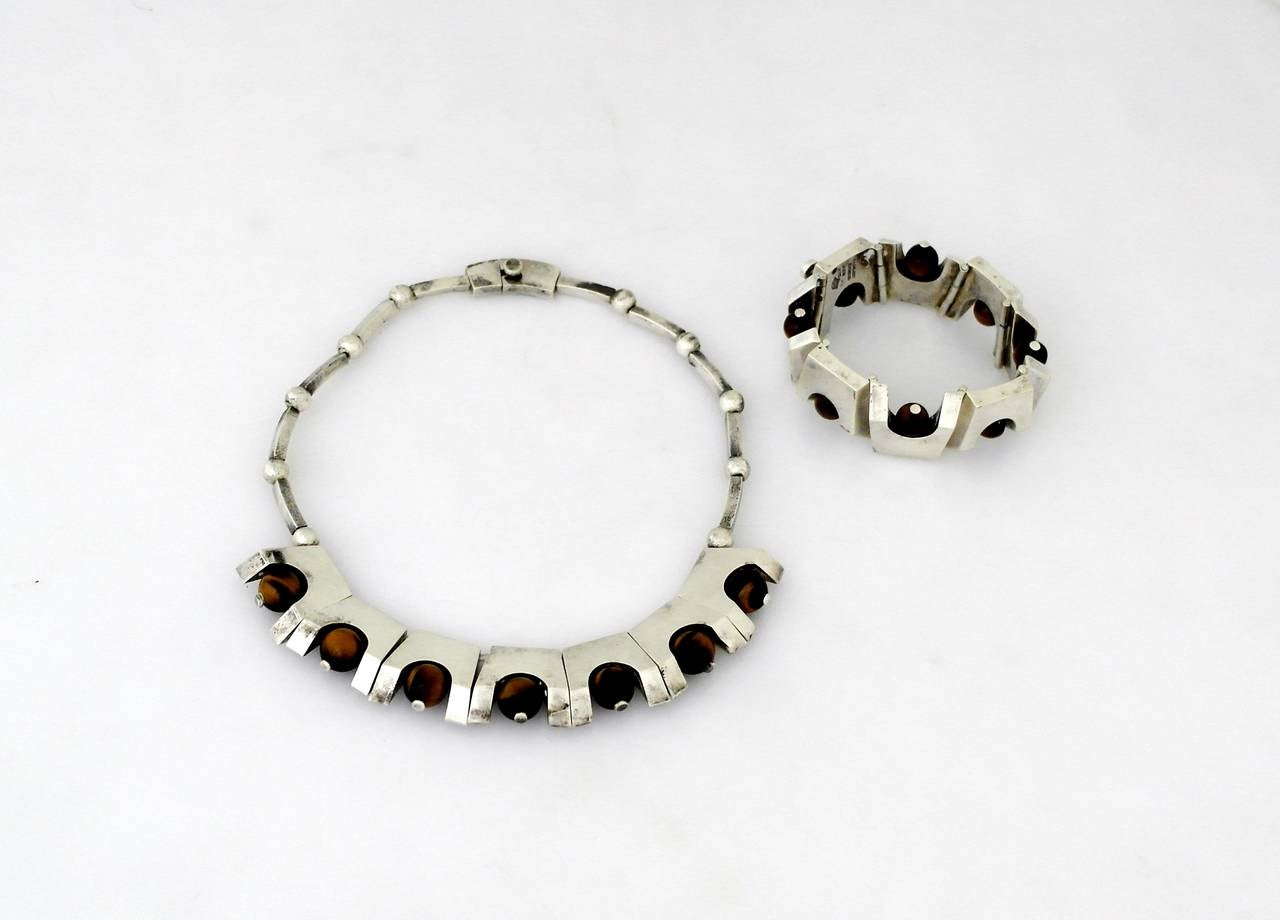 Being offered are a circa 1967 .970 silver jewelry suite by Antonio Pineda; comprising trapezoidal links with round tiger's eye stones set on both the necklace & bracelet.  Each 'link' is cast and measures 1 inch by 1 inch.  Marked as illustrated.
