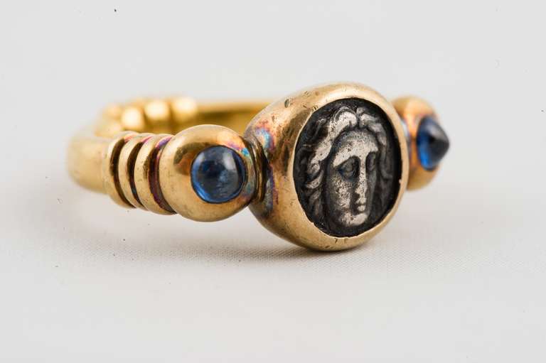 22K Yellow Gold Ring, bezel set with Roman coin (Satraps of Karia. Pixodaros AR Obol. Halikarnassos, 340-334 BC. Facing head of Apollo/Eight-rayed star) and two round cabochon blue sapphires weighing about .59 carat, on distressed looking shank with