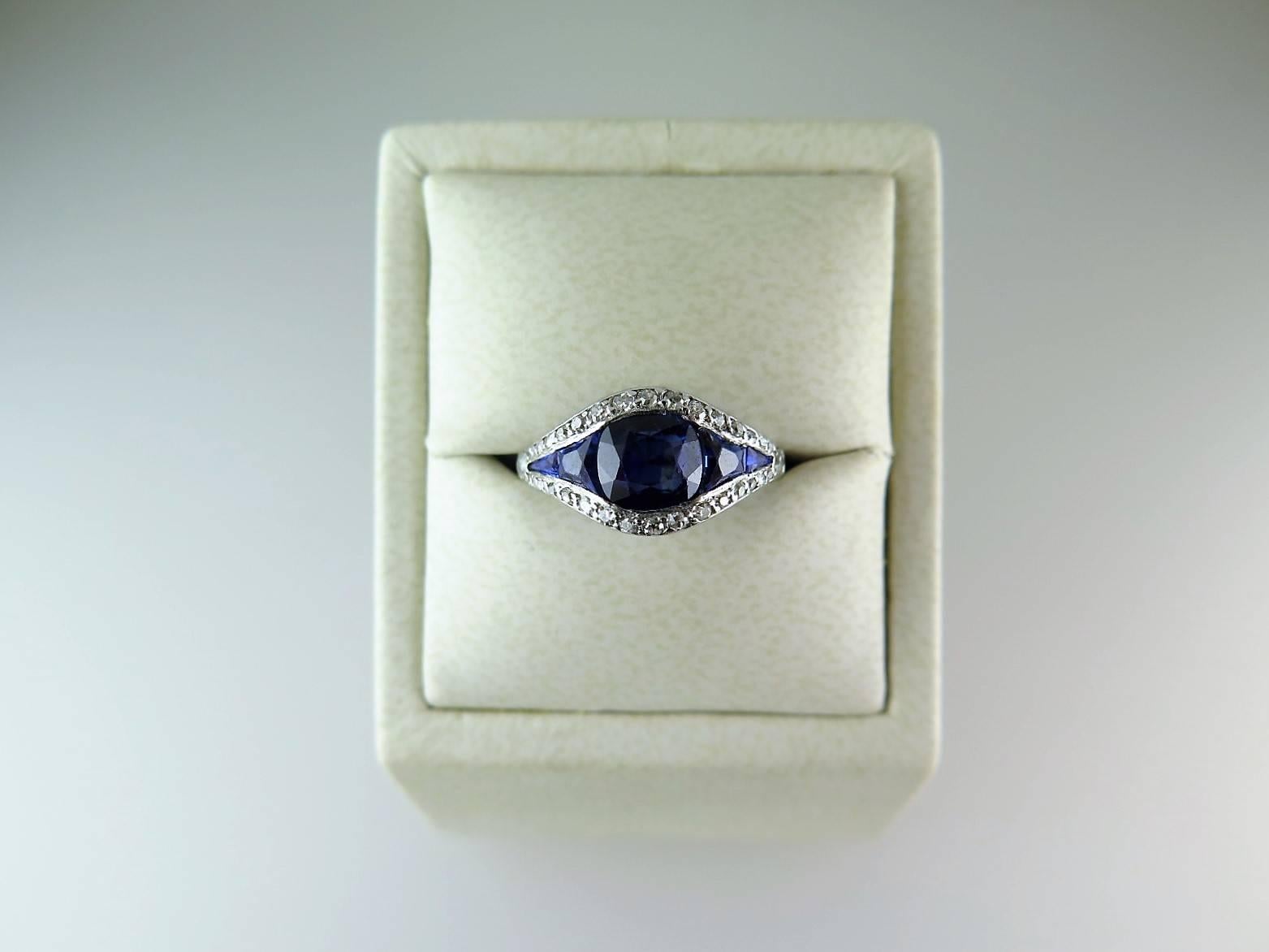 The central cushion-shaped sapphire calculated to weigh approx. 1.25cts with calibré-cut sapphire shoulders, edged in single and old mine-cut diamonds to the plain platinum mount, size M (US size 6 1/2). Can be resized to order.