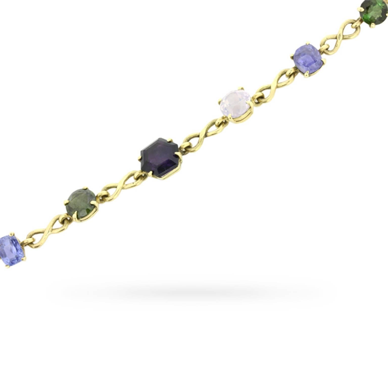 This vintage circa 1960s multi-gem bracelet features a colourful array of precious and semi-precious gemstones claw-set in open back mountings connected by infinity links in 18 carat yellow gold. The gemstones include sapphire, yellow sapphire,