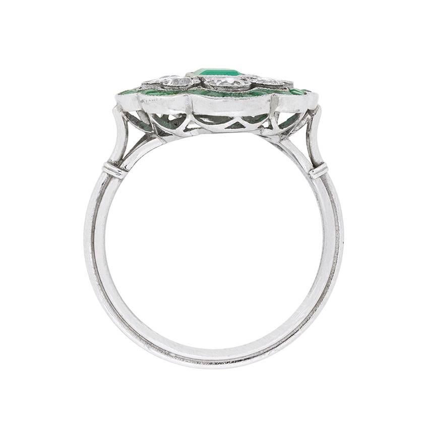 This lovely and floral cluster ring boasts a centre Emerald stone weighing 0.50 carat. It is grass green in colour, within a rub over setting. The stone is then encircled by old cut diamonds, confirming the rings age. The diamonds have a combined
