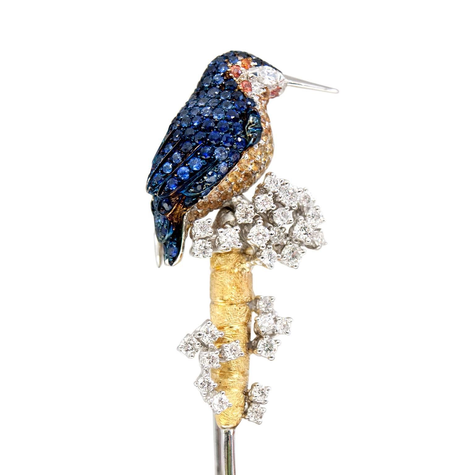 This is an elegant brooch with a kingfisher covered by many bright color stones such as sapphires. 
The eye of this intriguing river bird is a diamond navette cut.
The bird is perched on a flower in a lake, this flower is yellow gold and features