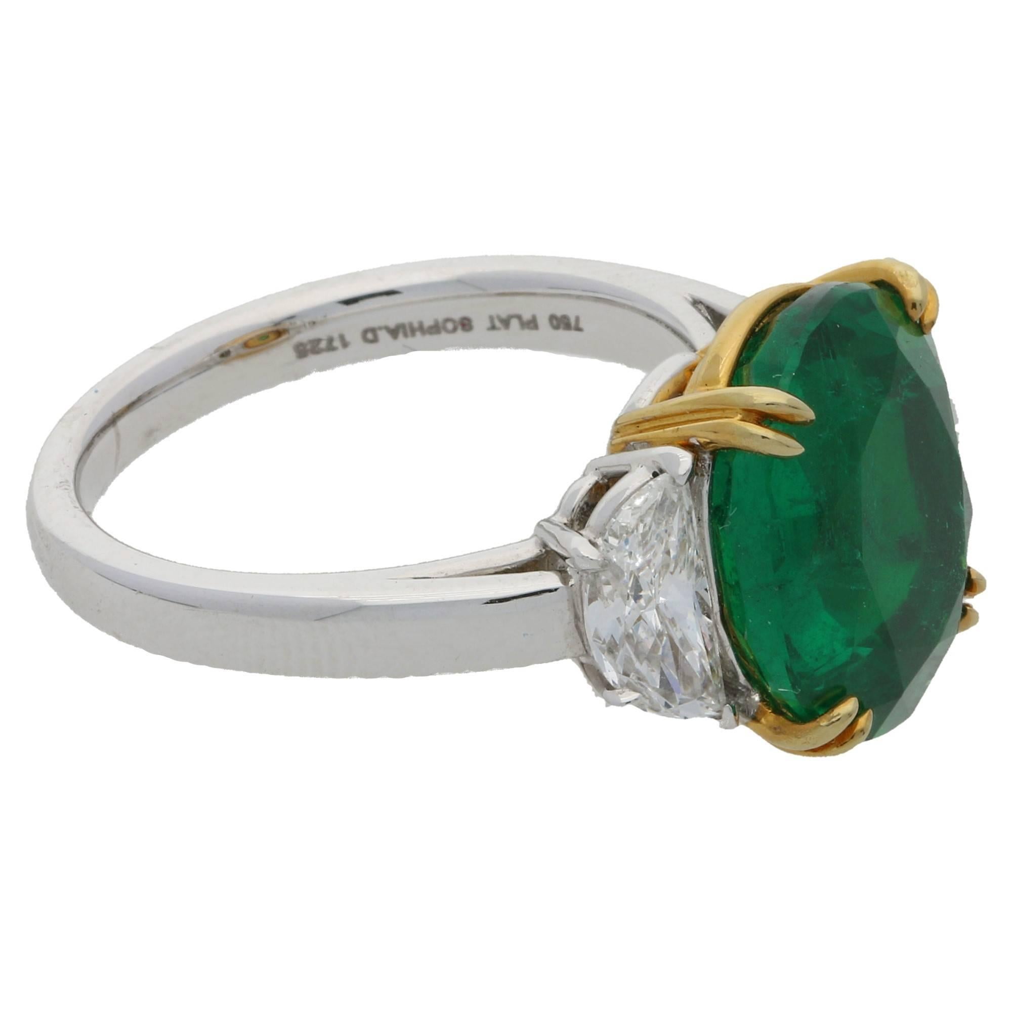 A three stone ring featuring an exquisite oval faceted Colombian emerald of a vibrant green colour with excellent conisistency of colour throughout the stone, set in a double four claw basket setting in 18ct yellow gold with a half moon cut diamond