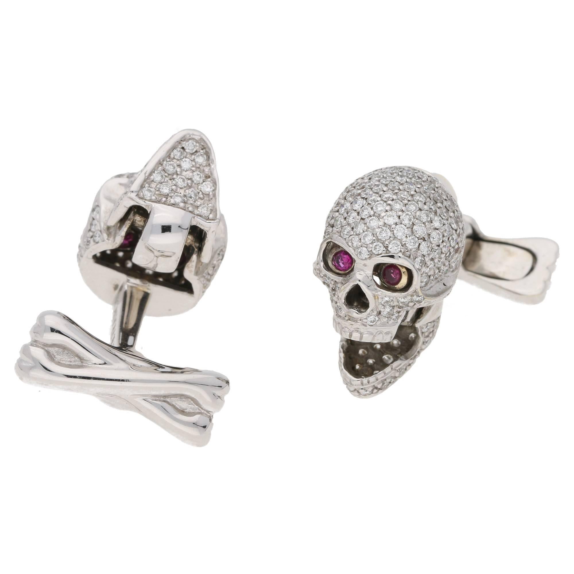 An outrageous pair of 18ct white gold skull and crossbones cufflinks. Each skull is fully pave set in round brilliant cut diamonds and has a hinged jaw that opens the mouth and reveals a hidden set of round faceted ruby eyes. On post and swivel