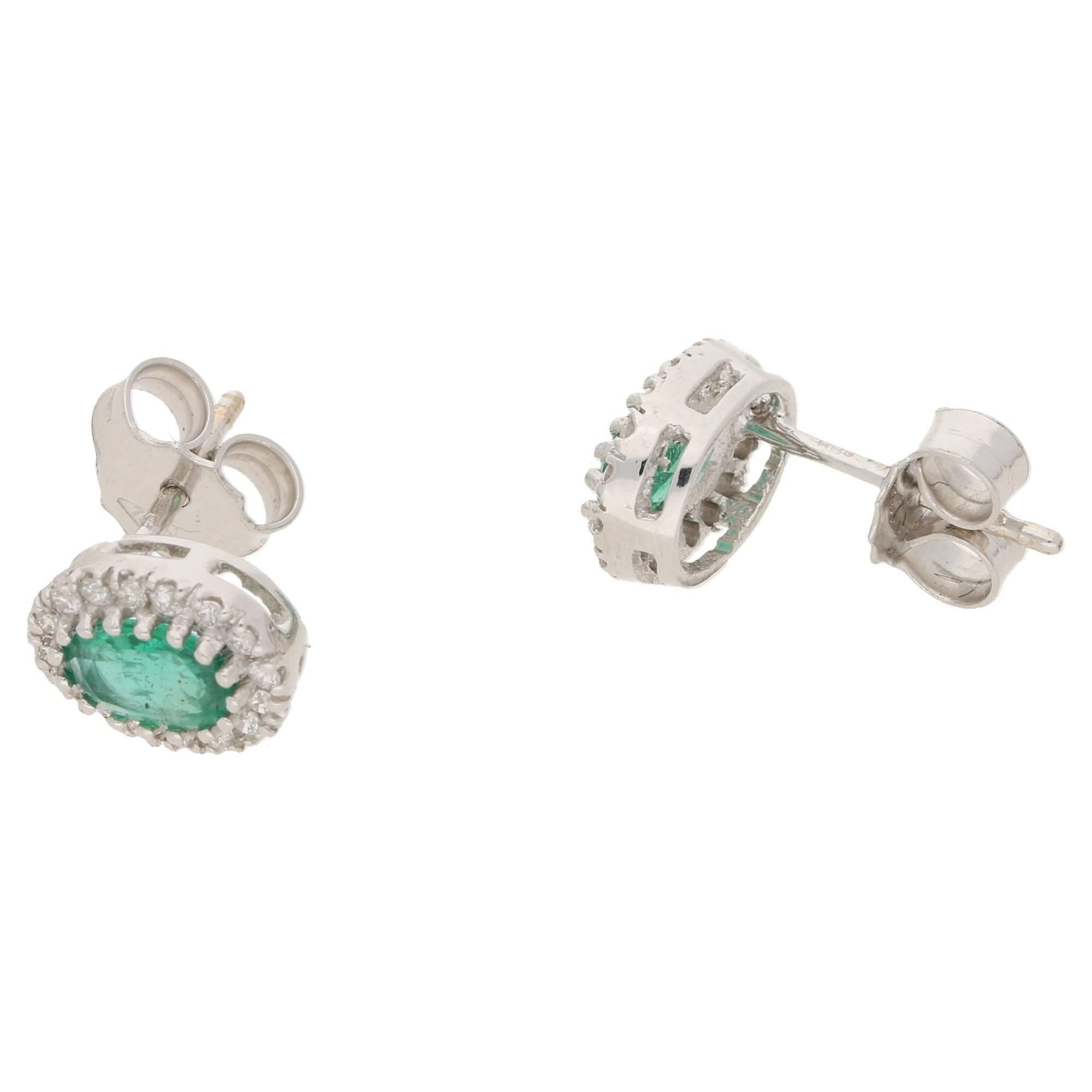 A pair of emerald and diamond cluster earrings. The earrings are formed of an oval cut emerald claw set to the centre, embellished by a single row of round brilliant cut diamonds. These earrings are made in 18ct white gold and have a post and scroll