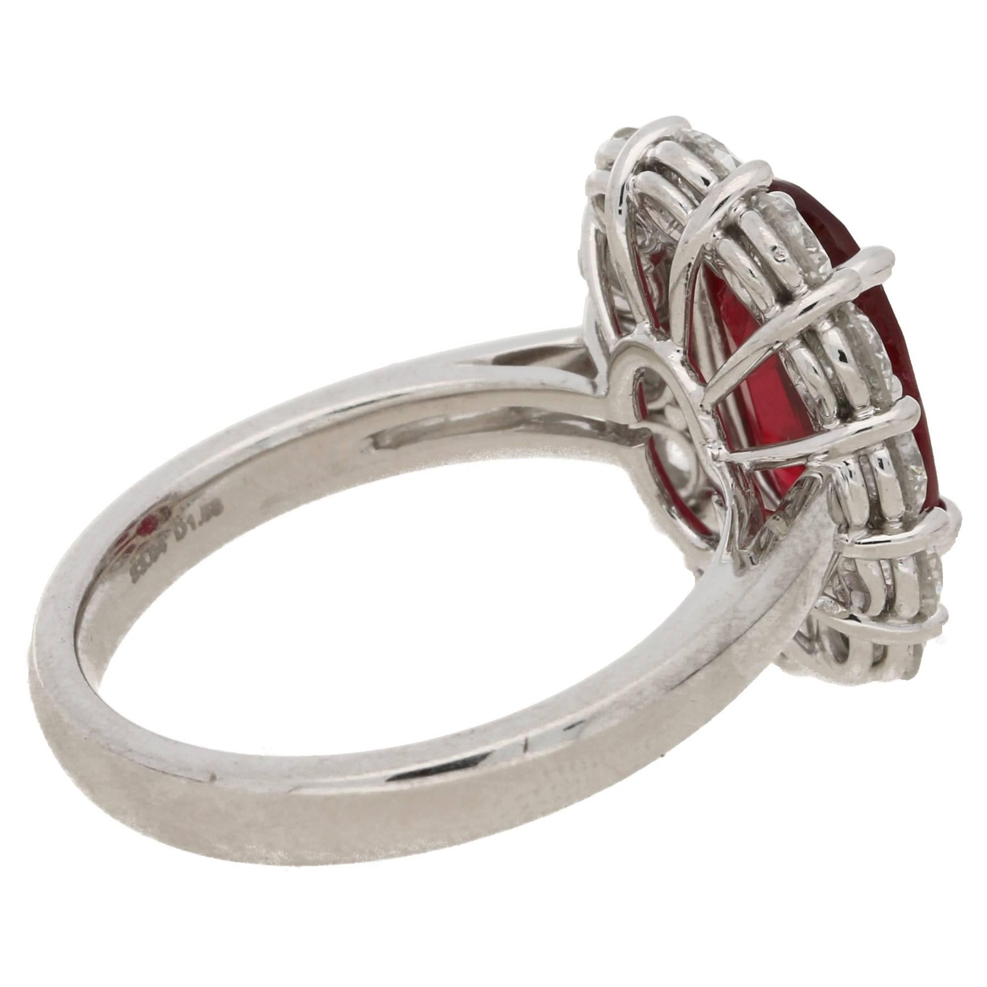 A classic oval ruby and diamond cluster ring set in platinum. The central long oval ruby is four-claw set in platinum amidst a border of twelve round brilliant cut diamonds mounted in a four-claw low impact setting, allowing the edge of the diamond