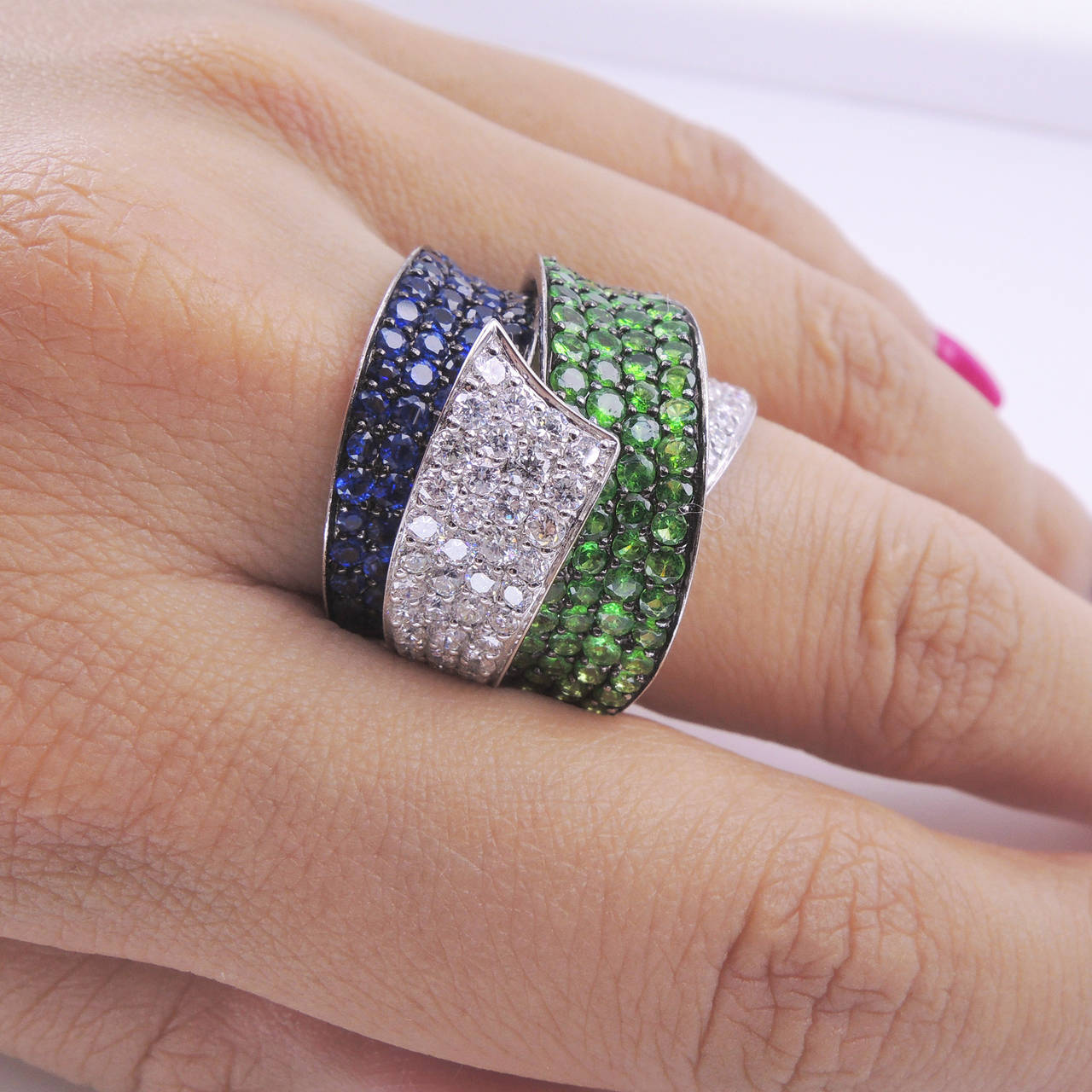 Stunning Diamond Tsavorite Sapphire Band Ring features an Intertwined design composed of 84 pave-set brilliant-cut diamonds=1.57ct, 63 sapphires=1.71ct and 84 tsavorite-1.99ct set in 18k white gold; Ring size 7; weight=18.02gm. Step out in Style and