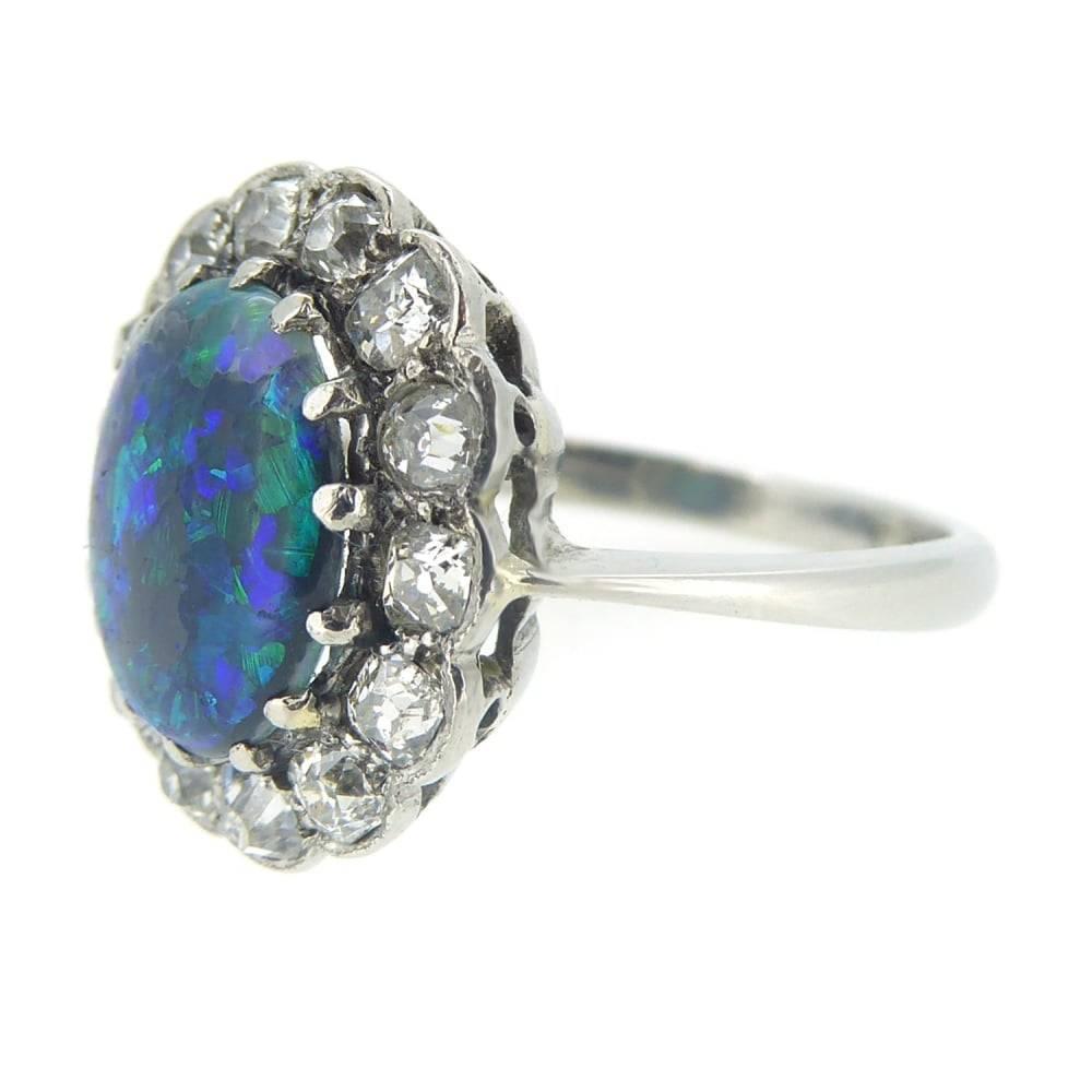 The spectacular green and blue/black play of colour in this cabochon cut opal is perfectly displayed within a border of old cut diamonds creating a dramatic and eye-catching cocktail ring.  The opal is supported by white claws and raised very