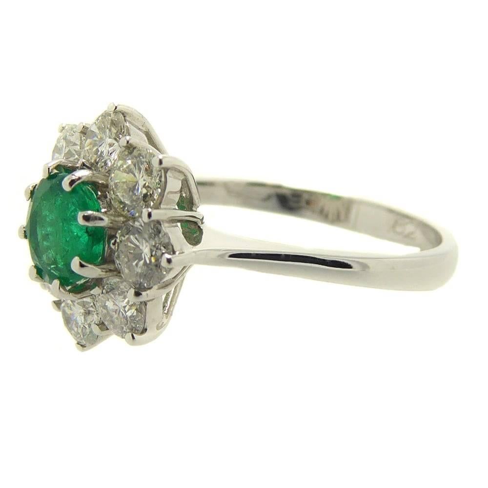 Classic style emerald and diamond cluster ring set to the centre with a round emerald of good strong green colour to a surround of eight very nice diamonds all claw set in white metal on a basket style mount.  Knife edge shoulders to a slight